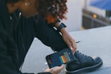 A person seated and holding a smartphone displaying a colorful sound or music application screen. The person is wearing a black outfit including black lace-up boots and a black wristwatch. The background appears to be an outdoor setting with a concrete or rooftop surface, and out-of-focus elements.