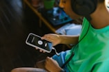 A person wearing large headphones is seated, holding a smartphone with a music app open. The screen displays a play/pause button and track information. The individual is casually dressed in a bright green shirt. A wooden floor and some potted plants are visible in the background.