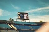 A large stadium with empty blue seats, a central video screen displaying a football player wearing a white jersey with the name Léo Ortiz and number 3. The sky is a bright blue with some clouds visible.