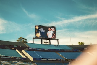 A large stadium with empty blue seats, a central video screen displaying a football player wearing a white jersey with the name Léo Ortiz and number 3. The sky is a bright blue with some clouds visible.