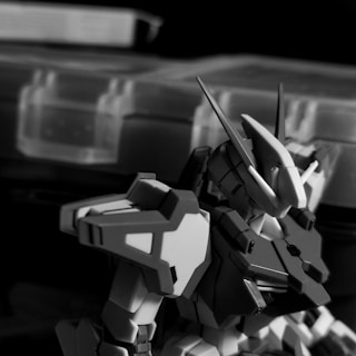 A detailed view of a model robot or mech with angular armor plating and a sleek design. The background features blurred storage containers, creating a sense of focus on the mechanical figure. The image is in black and white, highlighting the contours and details of the design.