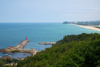 A coastal landscape features a wide expanse of blue ocean meeting the horizon under a partly cloudy sky. A long, narrow breakwater extends into the sea with a prominent red lighthouse near the end. To the right, a sandy beach stretches along the shoreline, bordered by lush, green forested areas. Buildings are visible in the distance along the coast.