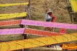 A person wearing a hat stands in front of several colorful fabrics stretched out on frames, likely for drying or inspection. The fabrics feature vibrant patterns in shades of yellow, orange, and pink. The setting appears outdoors on grassy terrain.