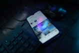 A smartphone is placed on a dark desk showing a social media interface, with visible icons and a grid of images on the screen. Nearby, there is a wireless mouse and part of a black computer keyboard. The lighting is low, highlighting blue and purple hues reflecting off the devices.