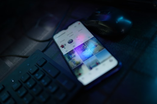 A smartphone is placed on a dark desk showing a social media interface, with visible icons and a grid of images on the screen. Nearby, there is a wireless mouse and part of a black computer keyboard. The lighting is low, highlighting blue and purple hues reflecting off the devices.