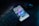 A smartphone is placed on a dark desk showing a social media interface, with visible icons and a grid of images on the screen. Nearby, there is a wireless mouse and part of a black computer keyboard. The lighting is low, highlighting blue and purple hues reflecting off the devices.