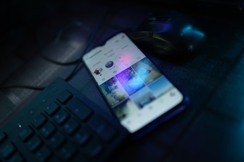 A smartphone is placed on a dark desk showing a social media interface, with visible icons and a grid of images on the screen. Nearby, there is a wireless mouse and part of a black computer keyboard. The lighting is low, highlighting blue and purple hues reflecting off the devices.