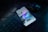 A smartphone is placed on a dark desk showing a social media interface, with visible icons and a grid of images on the screen. Nearby, there is a wireless mouse and part of a black computer keyboard. The lighting is low, highlighting blue and purple hues reflecting off the devices.