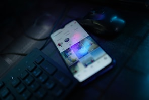 A smartphone is placed on a dark desk showing a social media interface, with visible icons and a grid of images on the screen. Nearby, there is a wireless mouse and part of a black computer keyboard. The lighting is low, highlighting blue and purple hues reflecting off the devices.