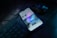 A smartphone is placed on a dark desk showing a social media interface, with visible icons and a grid of images on the screen. Nearby, there is a wireless mouse and part of a black computer keyboard. The lighting is low, highlighting blue and purple hues reflecting off the devices.