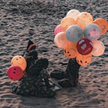 Two children sitting on a sandy beach, engaged in an activity. They are surrounded by colorful balloons with smiley faces and various designs, including the word 'LOVE' printed on some. The children wear patterned clothing and appear to be sitting closely together.