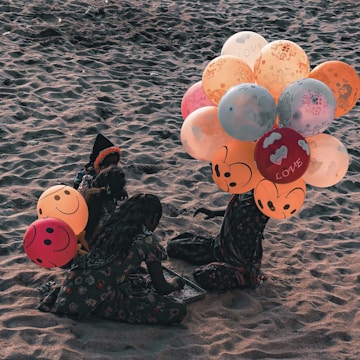 Two children sitting on a sandy beach, engaged in an activity. They are surrounded by colorful balloons with smiley faces and various designs, including the word 'LOVE' printed on some. The children wear patterned clothing and appear to be sitting closely together.