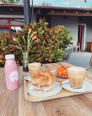 A wooden table is set with various breakfast items including a pink bottled smoothie, a vase with pink flowers, and a tray containing two seeded bagels filled with ham and greens, alongside two frothy lattes in clear glasses. In the background, vibrant green foliage and a modern building with red trim are visible.