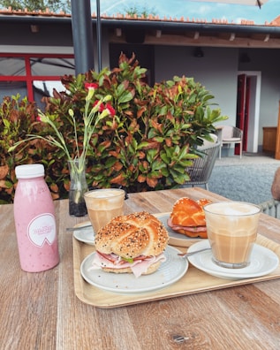 A wooden table is set with various breakfast items including a pink bottled smoothie, a vase with pink flowers, and a tray containing two seeded bagels filled with ham and greens, alongside two frothy lattes in clear glasses. In the background, vibrant green foliage and a modern building with red trim are visible.