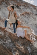 Two women are positioned on rugged, rocky terrain. One stands confidently, wearing a fringed vest and jeans, looking across the landscape. The other is seated, dressed in a white dress, holding a large dreamcatcher with feathers. The background features dramatic, textured rock formations.