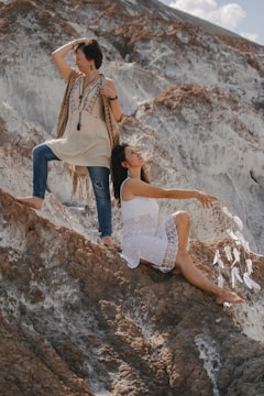 Two women are positioned on rugged, rocky terrain. One stands confidently, wearing a fringed vest and jeans, looking across the landscape. The other is seated, dressed in a white dress, holding a large dreamcatcher with feathers. The background features dramatic, textured rock formations.