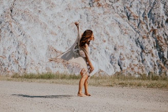 A person dances gracefully on a gravel path, wearing a beige fringed shawl and a patterned dress. The background features a textured rocky cliff with sparse grassy patches at the base.