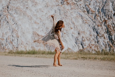 A person dances gracefully on a gravel path, wearing a beige fringed shawl and a patterned dress. The background features a textured rocky cliff with sparse grassy patches at the base.
