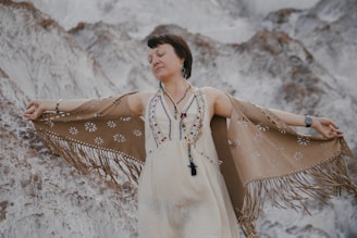A person with closed eyes is wearing a white dress adorned with colorful beads and a fringed tan shawl. They are standing against a rocky or mottled background, with arms outstretched in a serene pose.