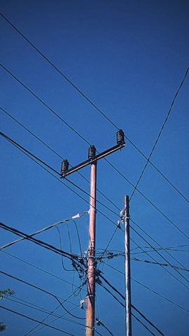 A network of power lines extends across a clear blue sky, supported by wooden poles with insulators. The wires intersect and connect at various points, creating a complex pattern against the backdrop.