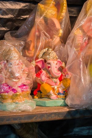 A collection of colorful Ganesha statues, partially wrapped in transparent plastic. The figures are ornate, featuring intricate details with golden crowns and jewelry, set against a wooden surface.