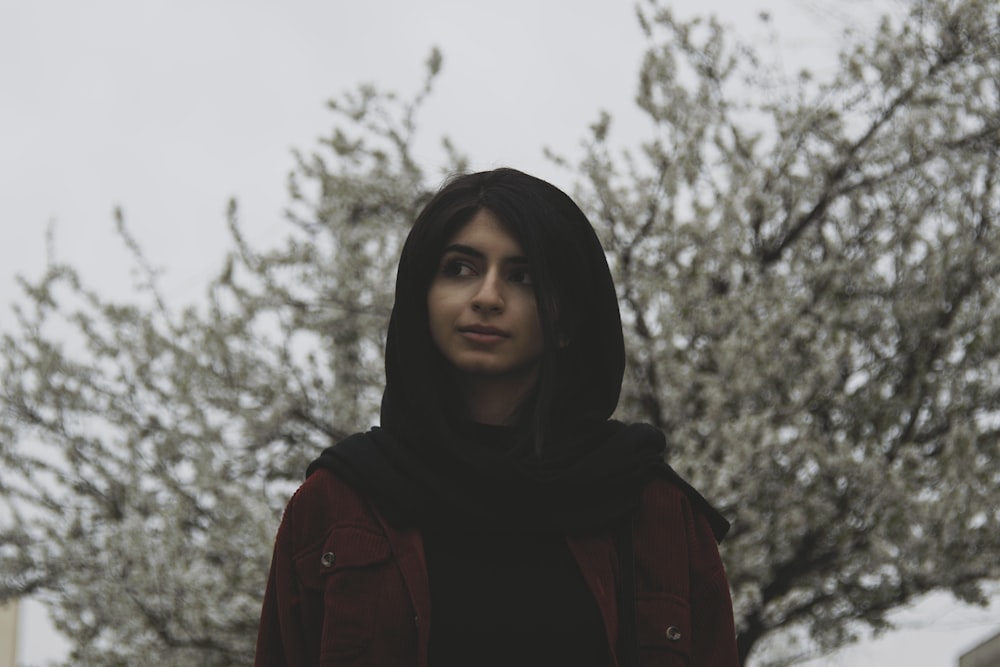 a woman wearing a black head scarf and a red jacket standing in front of a tree with snow