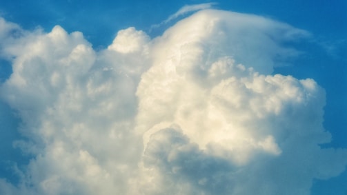A large, fluffy cumulus cloud fills the sky, illuminated by sunlight against a backdrop of bright blue. The cloud has a soft texture and appears expansive and towering, with various shades of white and gray highlighting its contours.