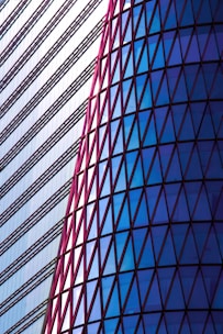 A striking architectural design featuring a modern building facade. The left side consists of horizontal lines with a minimalist style, while the right side displays a series of triangular glass panels in a spectrum of blues framed by red outlines.