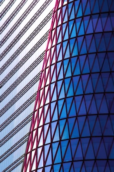 A striking architectural design featuring a modern building facade. The left side consists of horizontal lines with a minimalist style, while the right side displays a series of triangular glass panels in a spectrum of blues framed by red outlines.