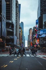 A busy urban street with pedestrians crossing at a crosswalk surrounded by tall buildings and bright electronic billboards. The sky is overcast, and the scene captures the hustle and bustle of a city environment.