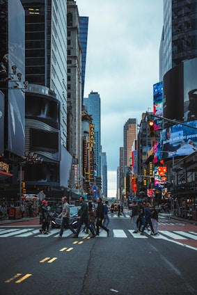 A busy urban street with pedestrians crossing at a crosswalk surrounded by tall buildings and bright electronic billboards. The sky is overcast, and the scene captures the hustle and bustle of a city environment.