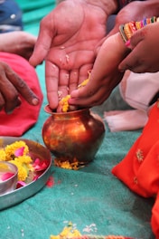 Several hands are involved in a traditional ritual, handling yellow flower petals and placing them in a copper vessel. A metal plate with marigold and rose petals is nearby. Bright, colorful clothing and ornaments, including bangles, are noticeable, suggesting a cultural or religious ceremony.