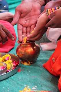 Several hands are involved in a traditional ritual, handling yellow flower petals and placing them in a copper vessel. A metal plate with marigold and rose petals is nearby. Bright, colorful clothing and ornaments, including bangles, are noticeable, suggesting a cultural or religious ceremony.