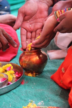 Several hands are involved in a traditional ritual, handling yellow flower petals and placing them in a copper vessel. A metal plate with marigold and rose petals is nearby. Bright, colorful clothing and ornaments, including bangles, are noticeable, suggesting a cultural or religious ceremony.