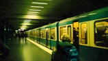 A metro train is stationed at an underground platform. Several people are visible, some standing inside the train while others are walking along the platform. The lighting is dim, and the scene appears to have a greenish tint.