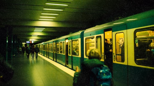 A metro train is stationed at an underground platform. Several people are visible, some standing inside the train while others are walking along the platform. The lighting is dim, and the scene appears to have a greenish tint.