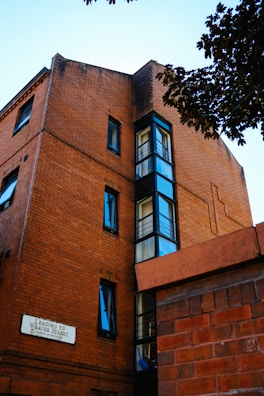 A brick building with multiple windows, featuring a sign that reads 'Leading to Weaver Street'. The architecture highlights a blend of traditional brickwork and modern glass elements. Trees and foliage partially frame the scene, adding a natural contrast.