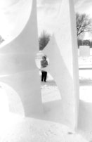 A person stands behind an abstract snow sculpture with angular shapes and openings. The scene is set outdoors on a snowy day, with bare trees and a house visible in the background. The sculpture has a modern, geometric design.