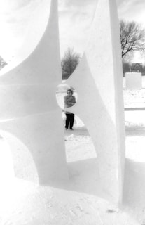 A person stands behind an abstract snow sculpture with angular shapes and openings. The scene is set outdoors on a snowy day, with bare trees and a house visible in the background. The sculpture has a modern, geometric design.