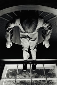 A child wearing a suit sits on a chair, viewed from above. The image is in black and white, creating a dramatic and introspective atmosphere. The child's hands rest on their knees and the chair is situated near a window with glass panes that reveal foliage outside.