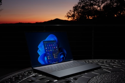 A laptop sits on a detailed metal table with a dark background. The screen is brightly lit, displaying a desktop interface with a blue-themed background. In the distance, a vivid sunset illuminates the sky with orange and pink hues, silhouetting trees and a hill.