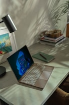 A cozy workspace featuring a laptop on a sleek, gray desk. The screen displays a modern interface with vivid colors. Nearby, there's a closed green notebook with a pen resting on top. A stylish desk lamp and a stack of books with a wooden decorative object are present. Soft shadows from nearby plants create a calming atmosphere.