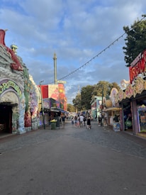 A pathway in an amusement park lined with colorful attractions and buildings. The buildings are adorned with vibrant paintings and sculptures, including a dragon and various fantasy-themed figures. Visitors, including families and groups of people, walk along the cobblestone path enjoying the park. The sky is partly cloudy with patches of blue visible.
