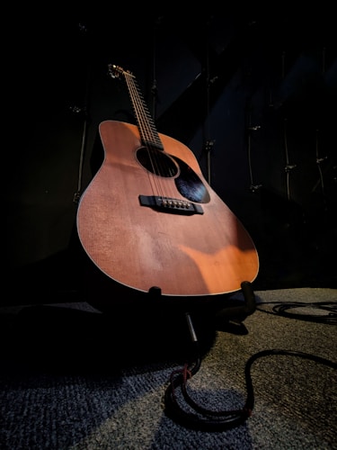 An acoustic guitar is positioned on a stand in a dimly lit room. The spotlight highlights the wooden texture and curves of the guitar. A cable is coiled near the base, suggesting it's ready for use or connection. The background is dark, enhancing the focus on the instrument.