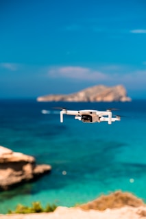 A small white drone hovers above a vibrant turquoise ocean with a distant rocky island blurred in the background under a clear blue sky.