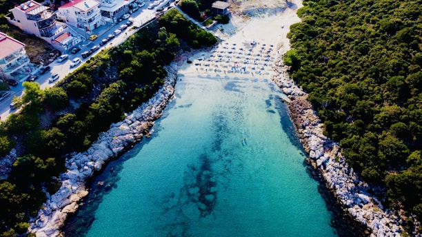 An aerial view of a picturesque cove with clear turquoise water surrounded by rocky and green vegetated cliffs. There is a beach area with neatly arranged umbrellas and sunbeds, suggesting a popular spot for relaxation and swimming. Buildings and a road with parked cars border the top of the image.
