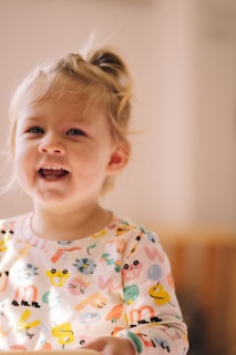 A young child with blonde hair is smiling brightly. The child is wearing a colorful shirt covered in various cartoon animals and shapes. The background is softly blurred, creating a warm and cozy atmosphere.