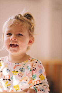 A young child with blonde hair is smiling brightly. The child is wearing a colorful shirt covered in various cartoon animals and shapes. The background is softly blurred, creating a warm and cozy atmosphere.