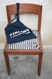 A wooden chair with a torn black and white striped cushion is positioned against a textured wall. A navy blue tote bag with the words 'Finland, A Tech Superpower' printed in white rests on the chair.