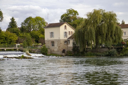 A serene riverside scene with a quaint two-story house featuring stone and cream-colored walls. The house is surrounded by lush greenery, including a large weeping willow tree. A gentle river flows in the foreground, with a small weir or water feature visible to the left. The background has dense green trees under a cloudy sky, adding to the tranquil setting.